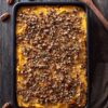 Overhead view of butternut squash casserole in a black baking dish, topped with chopped pecans, breadcrumbs, and herbs on a dark rustic wood table.