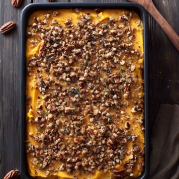 Overhead view of butternut squash casserole in a black baking dish, topped with chopped pecans, breadcrumbs, and herbs on a dark rustic wood table.