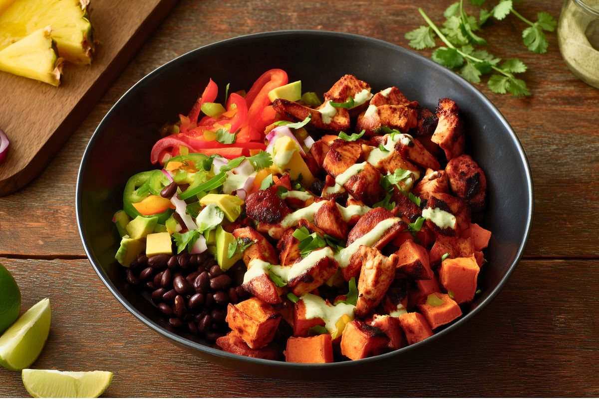 Caribbean chicken sweet potato bowl in a black bowl with jerk-seasoned chicken, roasted sweet potatoes, black beans, avocado, peppers, red onion, pineapple, cilantro, and creamy green sauce on a rustic wood table.