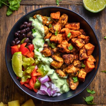 Overhead view of a Caribbean chicken sweet potato bowl with seasoned chicken, roasted sweet potatoes, black beans, red and green peppers, red onion, and creamy green sauce in a black bowl on dark rustic wood.