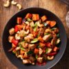 Overhead view of a cashew chicken sweet potato bowl with glazed chicken, roasted sweet potatoes, zucchini, red bell pepper, cashews, and sliced green onions in a black bowl on a rustic wood table.