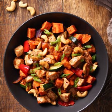 Overhead view of a cashew chicken sweet potato bowl with glazed chicken, roasted sweet potatoes, zucchini, red bell pepper, cashews, and sliced green onions in a black bowl on a rustic wood table.