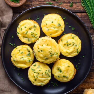 Overhead view of golden cheddar and chive egg bites on a black plate with fresh chives and sliced cheddar cheese on a rustic wood table.