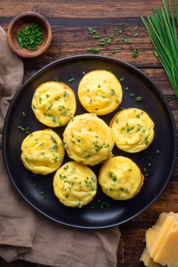 Overhead view of golden cheddar and chive egg bites on a black plate with fresh chives and sliced cheddar cheese on a rustic wood table.