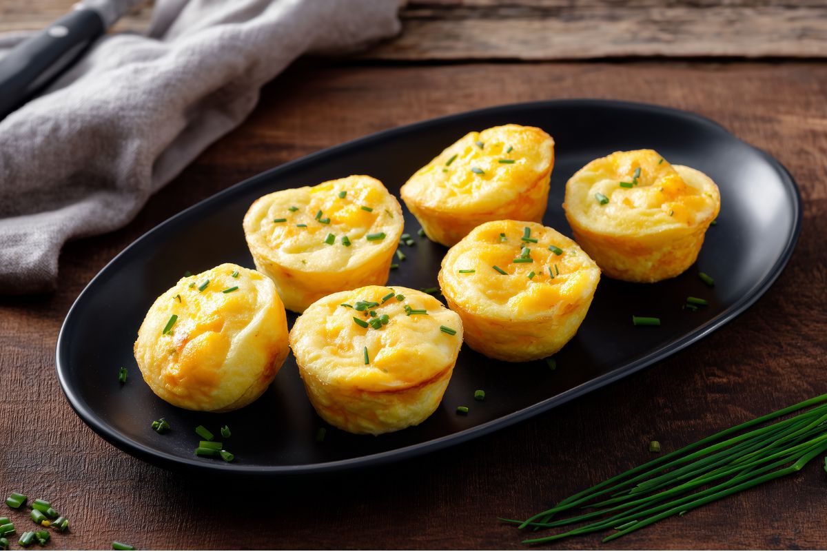 Golden cheddar and chive egg bites arranged on a black oval plate with fresh chives on a rustic wooden table.