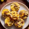Cheeseburger egg bites with browned ground beef, melted cheddar cheese, and chopped pickles arranged on a plate with whole dill pickles nearby.