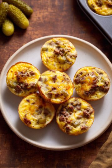 Cheeseburger egg bites with browned ground beef, melted cheddar cheese, and chopped pickles arranged on a plate with whole dill pickles nearby.