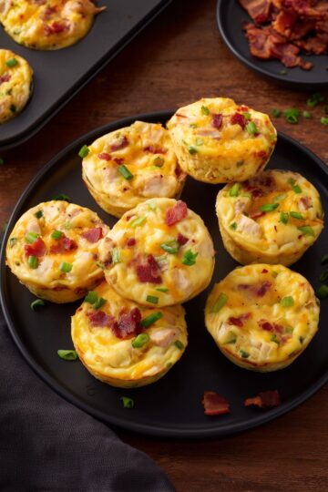 Overhead view of chicken bacon ranch egg bites topped with bacon, chicken, melted cheese, and green onions on a black plate, with a muffin pan and extra bacon nearby.