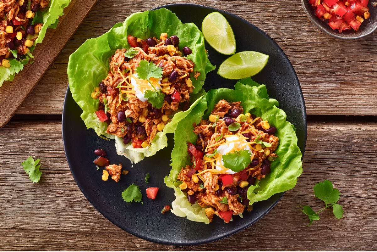 Chicken burrito lettuce wraps filled with seasoned chicken, black beans, corn, tomatoes, shredded cheese, creamy sauce, and cilantro on a black plate over rustic wood.