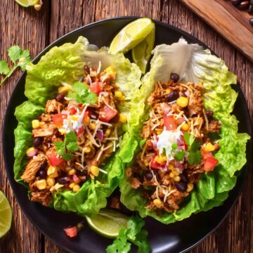 Overhead view of chicken burrito lettuce wraps with seasoned chicken, black beans, corn, tomatoes, shredded cheese, creamy sauce, and fresh cilantro on a black plate.