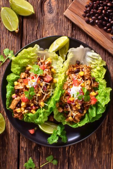 Overhead view of chicken burrito lettuce wraps with seasoned chicken, black beans, corn, tomatoes, shredded cheese, creamy sauce, and fresh cilantro on a black plate.