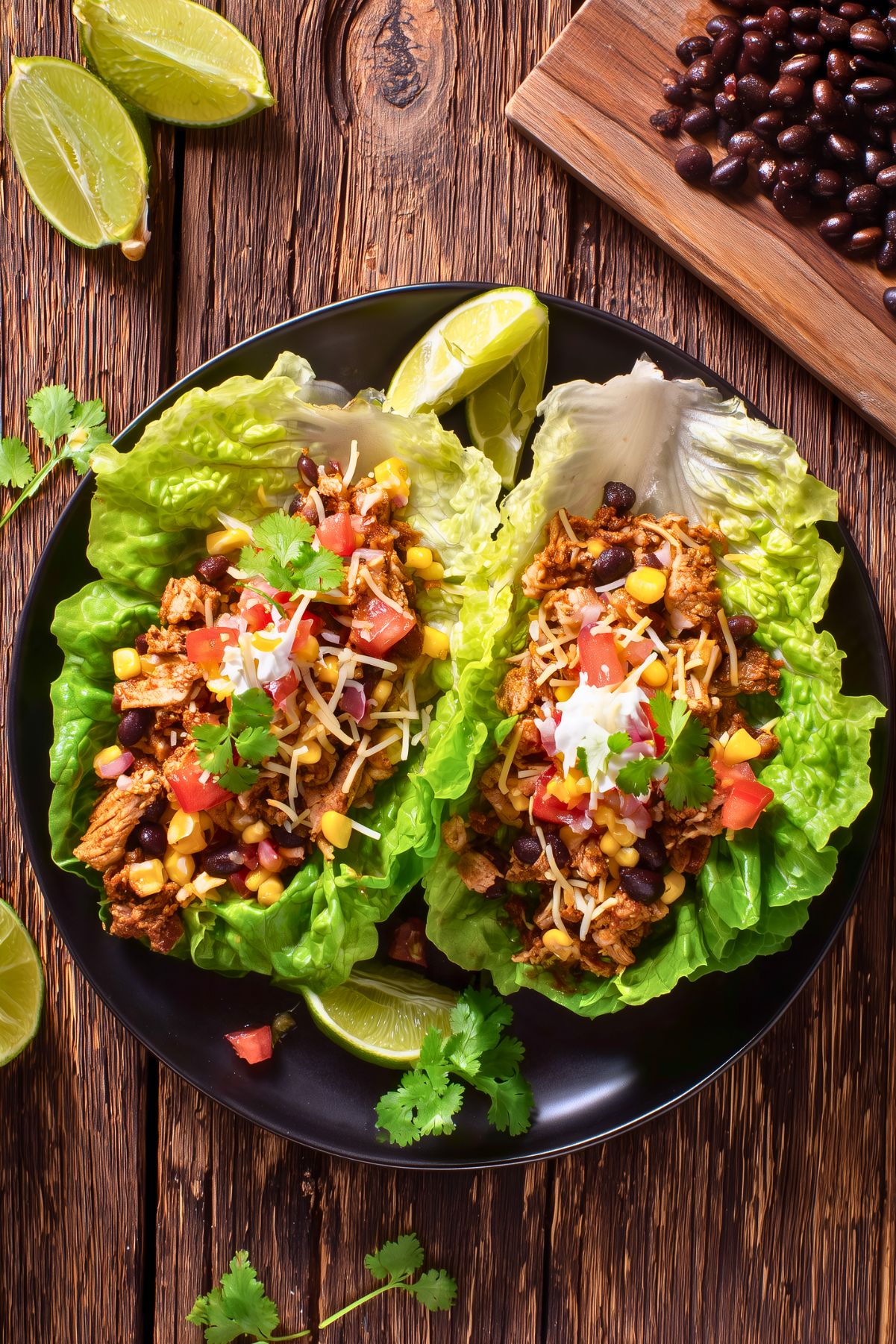 Overhead view of chicken burrito lettuce wraps with seasoned chicken, black beans, corn, tomatoes, shredded cheese, creamy sauce, and fresh cilantro on a black plate.