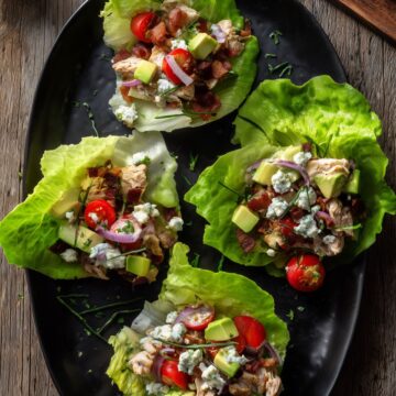 Overhead view of Chicken Cobb lettuce wraps with chicken, bacon, avocado, tomatoes, red onion and blue cheese served on a black platter over rustic wood.