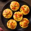 Overhead view of six chicken enchilada egg bites with melted cheese, red pepper, and green onions on a black plate over rustic wood.