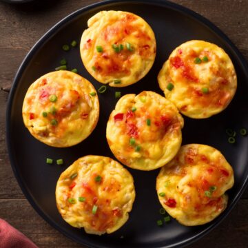 Overhead view of six chicken enchilada egg bites with melted cheese, red pepper, and green onions on a black plate over rustic wood.
