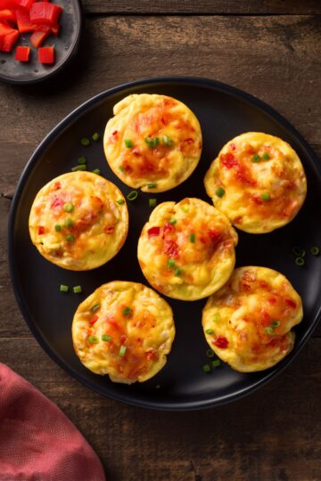 Overhead view of six chicken enchilada egg bites with melted cheese, red pepper, and green onions on a black plate over rustic wood.