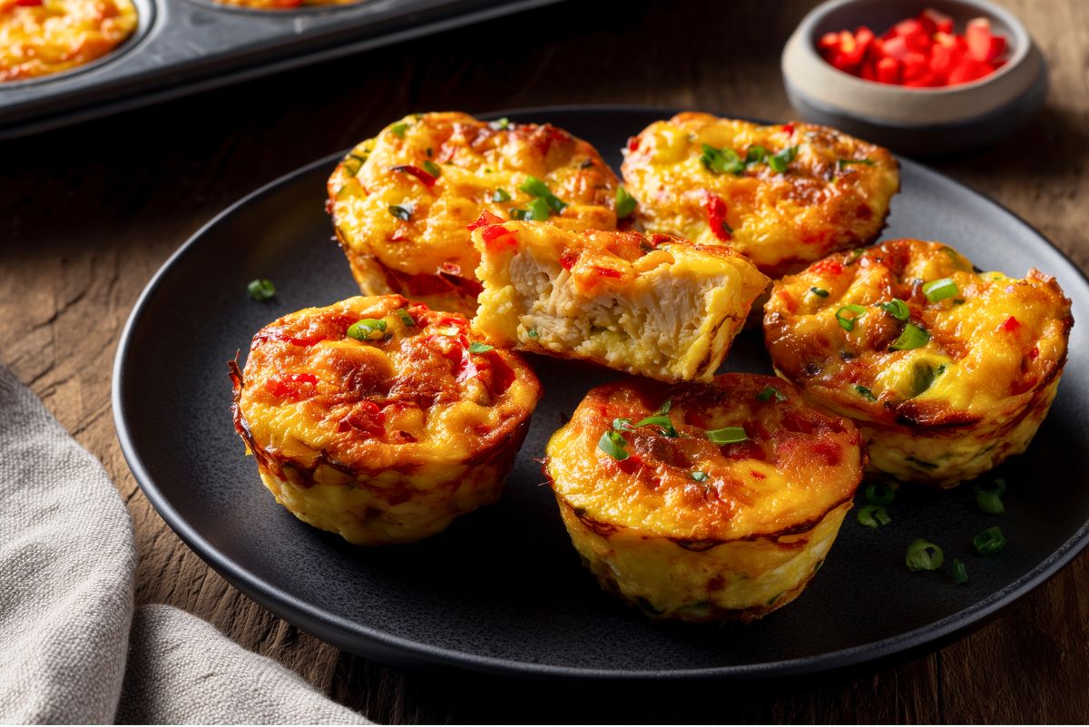 Chicken enchilada egg bites on a black plate, topped with melted cheese, red pepper, and green onions, with one cut open to show the chicken filling.
