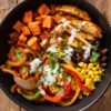 Overhead chicken fajita sweet potato bowl with seasoned chicken, roasted sweet potatoes, black beans, corn, sautéed red, yellow, and green peppers, onions, cilantro, and creamy sauce in a black bowl on rustic wood.