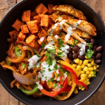 Overhead chicken fajita sweet potato bowl with seasoned chicken, roasted sweet potatoes, black beans, corn, sautéed red, yellow, and green peppers, onions, cilantro, and creamy sauce in a black bowl on rustic wood.