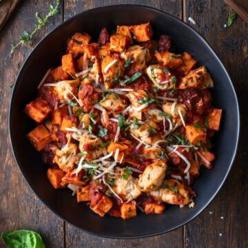 Overhead view of a Chicken Parmesan Sweet Potato Bowl with roasted sweet potato cubes, seasoned chicken, marinara sauce, shredded Parmesan and fresh basil in a black bowl.