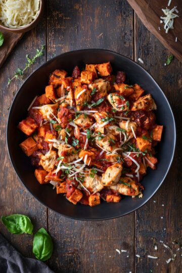 Overhead view of a Chicken Parmesan Sweet Potato Bowl with roasted sweet potato cubes, seasoned chicken, marinara sauce, shredded Parmesan and fresh basil in a black bowl.