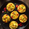 Six Chicken Taco Egg Bites with chicken, melted cheddar, red bell pepper, and green onions arranged on a matte black plate over a rustic wooden surface.