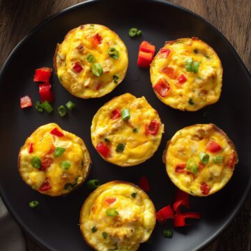 Six Chicken Taco Egg Bites with chicken, melted cheddar, red bell pepper, and green onions arranged on a matte black plate over a rustic wooden surface.