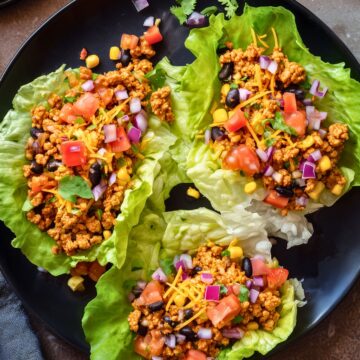 Overhead view of three chicken taco lettuce wraps on a black plate, filled with seasoned chicken, black beans, corn, tomatoes, red onion, shredded cheese, and cilantro.