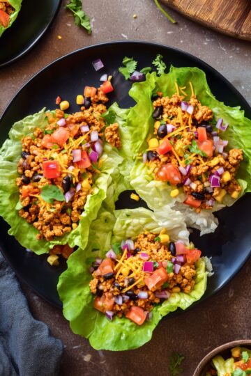 Overhead view of three chicken taco lettuce wraps on a black plate, filled with seasoned chicken, black beans, corn, tomatoes, red onion, shredded cheese, and cilantro.