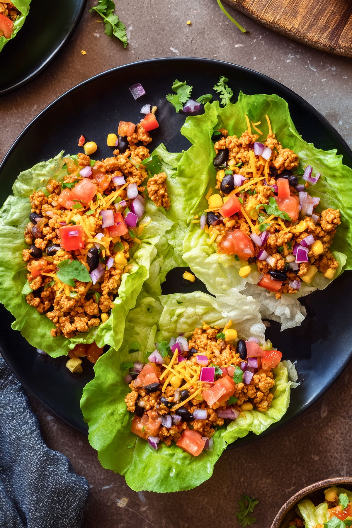 Overhead view of three chicken taco lettuce wraps on a black plate, filled with seasoned chicken, black beans, corn, tomatoes, red onion, shredded cheese, and cilantro.