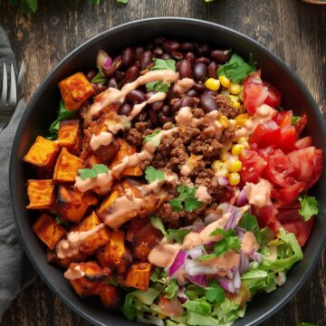 Overhead view of a Chipotle Beef Sweet Potato Bowl with roasted sweet potatoes, seasoned ground beef, black beans, corn, tomatoes, lettuce, red onion, cilantro, and creamy chipotle sauce in a black bowl on a rustic wood table.