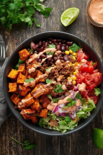Overhead view of a Chipotle Beef Sweet Potato Bowl with roasted sweet potatoes, seasoned ground beef, black beans, corn, tomatoes, lettuce, red onion, cilantro, and creamy chipotle sauce in a black bowl on a rustic wood table.