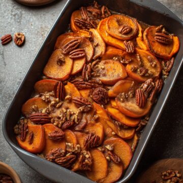 Cinnamon Apple Sweet Potato Casserole with tender sliced sweet potatoes and apples coated in a cinnamon maple glaze and topped with pecans, served in a dark baking dish with fresh apples nearby.