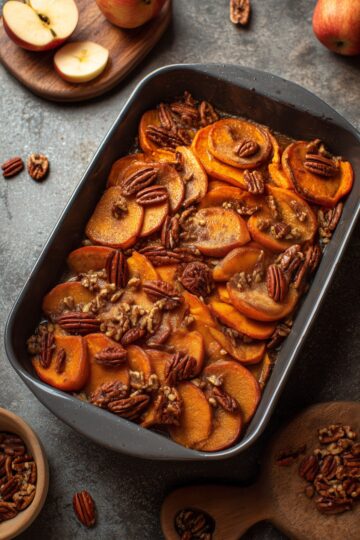 Cinnamon Apple Sweet Potato Casserole with tender sliced sweet potatoes and apples coated in a cinnamon maple glaze and topped with pecans, served in a dark baking dish with fresh apples nearby.