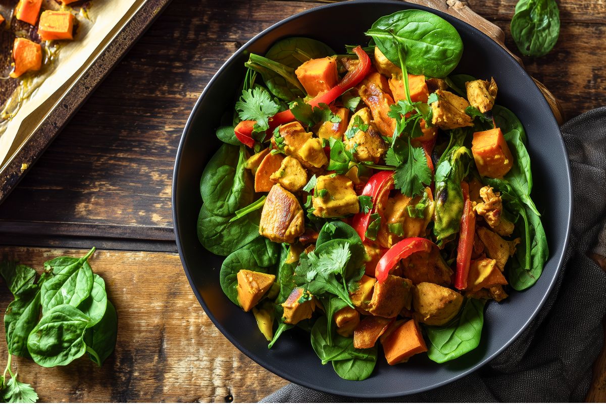 Overhead view of a coconut curry chicken sweet potato bowl with roasted sweet potatoes, seasoned chicken, red bell pepper, fresh spinach, cilantro, and golden curry sauce in a black bowl.