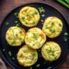 Overhead view of six cottage cheese egg bites with cheddar and green onions on a black plate over a rustic wood table.