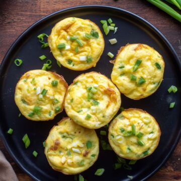 Overhead view of six cottage cheese egg bites with cheddar and green onions on a black plate over a rustic wood table.