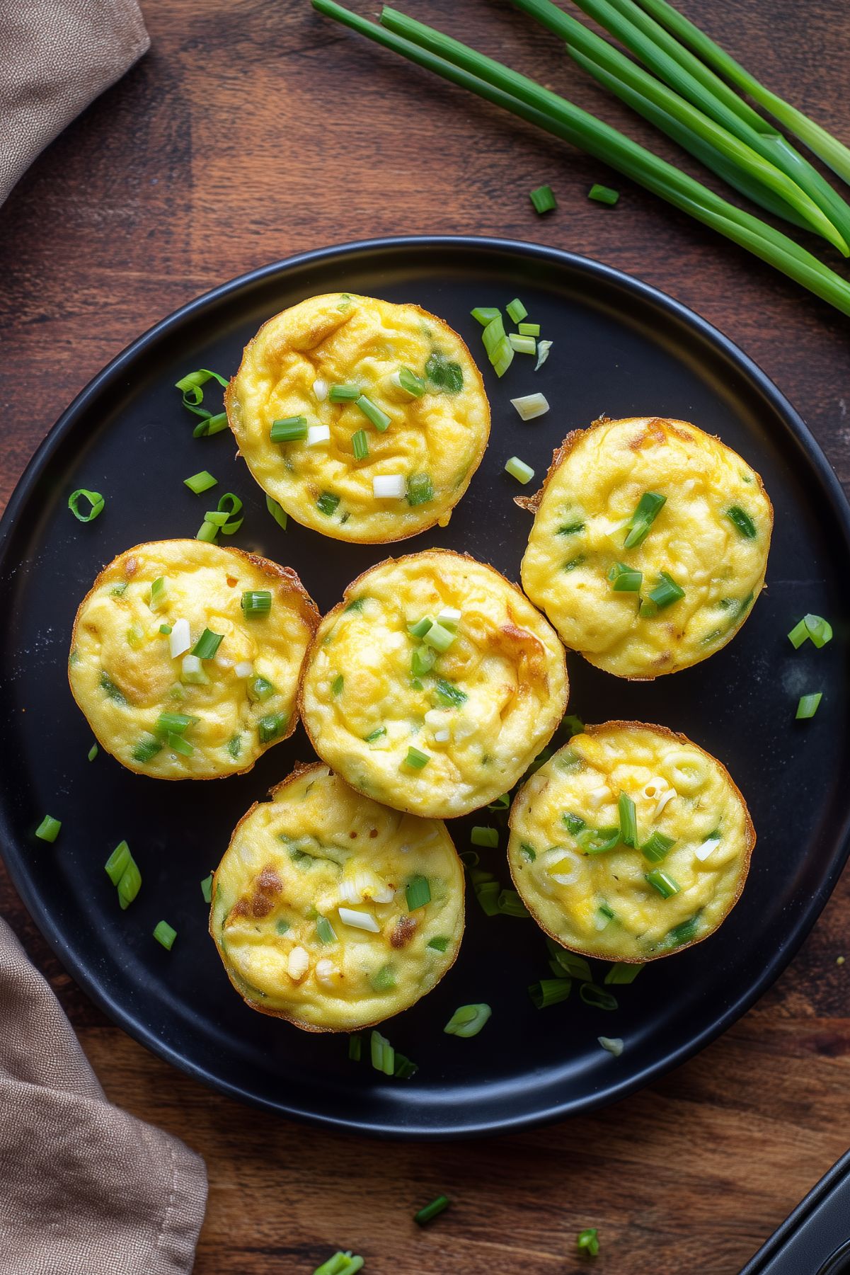 Overhead view of six cottage cheese egg bites with cheddar and green onions on a black plate over a rustic wood table.