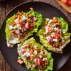 Overhead view of three Cottage Cheese and Turkey Lettuce Wraps on a black plate, topped with seasoned ground turkey, cottage cheese sauce, tomatoes, red onion, cucumber, and fresh parsley.