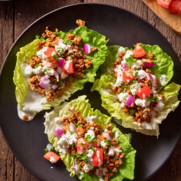 Overhead view of three Cottage Cheese and Turkey Lettuce Wraps on a black plate, topped with seasoned ground turkey, cottage cheese sauce, tomatoes, red onion, cucumber, and fresh parsley.