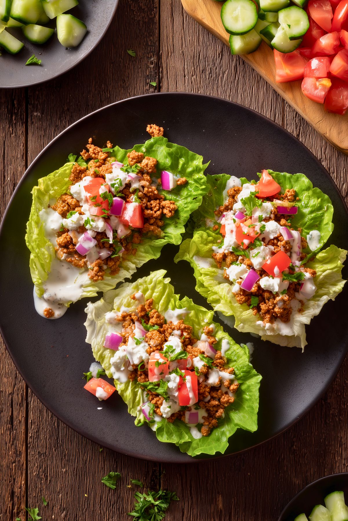 Overhead view of three Cottage Cheese and Turkey Lettuce Wraps on a black plate, topped with seasoned ground turkey, cottage cheese sauce, tomatoes, red onion, cucumber, and fresh parsley.