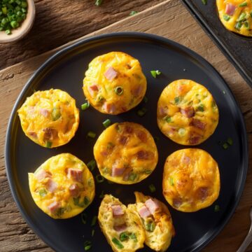 Overhead view of Denver omelet egg bites with ham, green peppers, cheddar cheese, and green onions arranged on a black plate over rustic wood, with more egg bites in a muffin pan.