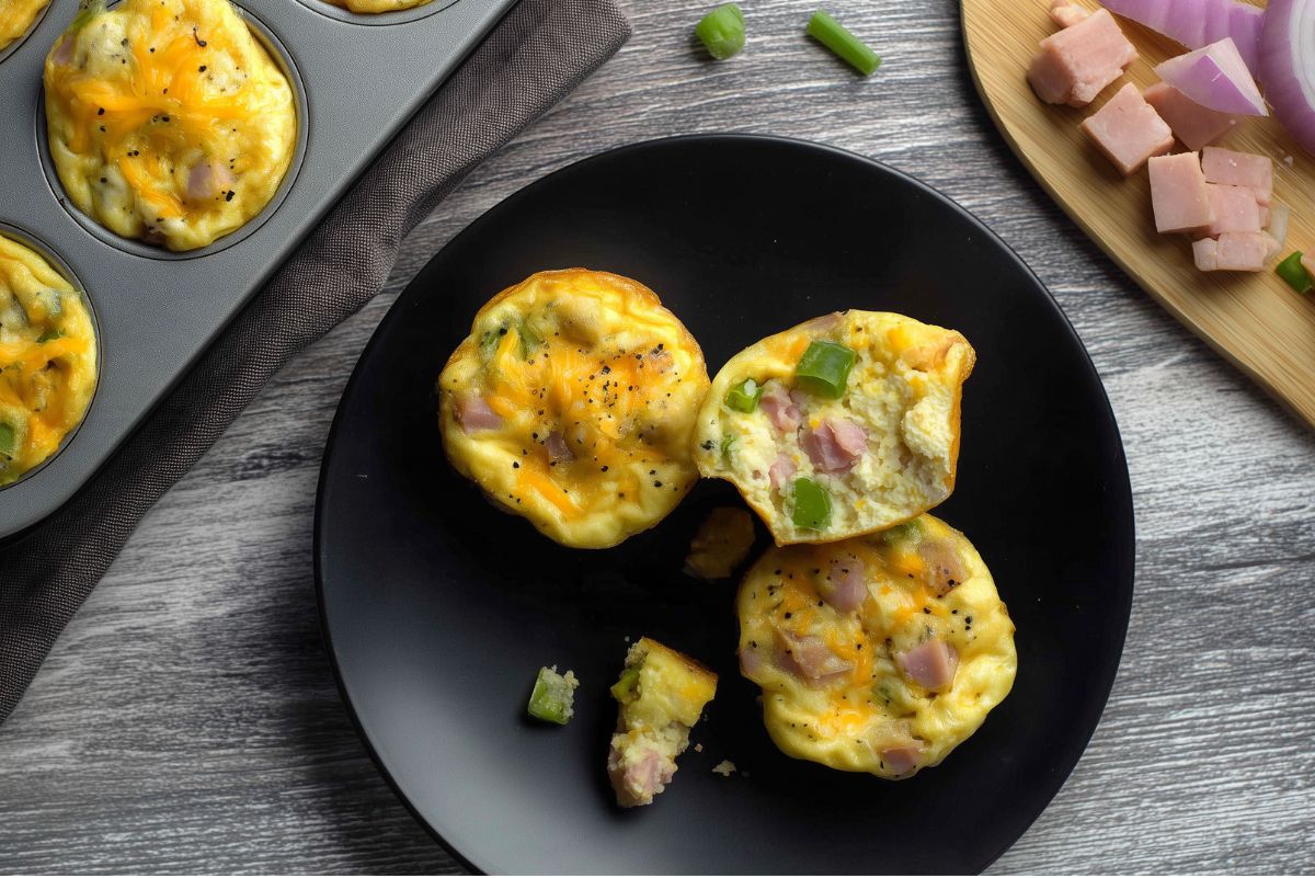 Denver omelet egg bites with diced ham, green bell pepper, and melted cheddar cheese on a black plate beside a muffin pan. One egg bite is cut open to show the soft egg filling.