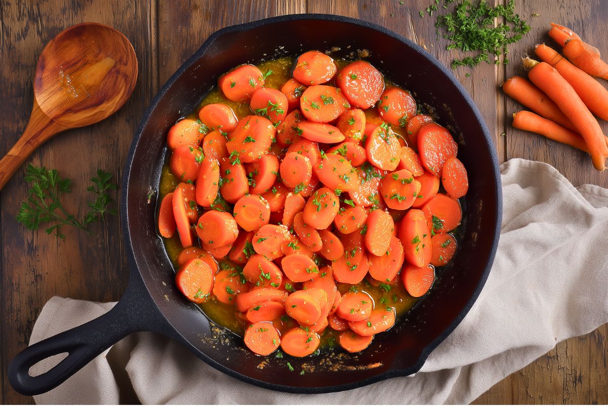 Sliced maple glazed carrots cooking in a black cast iron skillet with fresh parsley, surrounded by a wooden spoon and whole carrots on a rustic wood table.