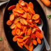 Easy maple glazed carrots with fresh parsley served on a black oval platter with a serving spoon, whole carrots, and a neutral linen napkin on a rustic wood table.