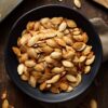 Overhead view of seasoned oven-roasted pumpkin seeds piled in a matte black bowl on a dark rustic wood table, with a baking sheet, wooden spoon, and scattered pumpkin seeds nearby.