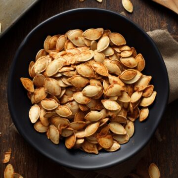 Overhead view of seasoned oven-roasted pumpkin seeds piled in a matte black bowl on a dark rustic wood table, with a baking sheet, wooden spoon, and scattered pumpkin seeds nearby.