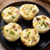 Five Egg White Turkey Bacon Bites with cheddar, turkey bacon, and green onions arranged on a black plate over a rustic wood table.