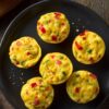 Six fajita vegetable egg bites with red and green bell peppers and corn arranged on a black plate over a rustic wood table.