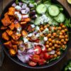 Overhead Greek chickpea sweet potato bowl with roasted sweet potatoes, spiced chickpeas, cucumber, cherry tomatoes, red onion, olives, leafy greens, feta, herbs, and creamy dressing in a black bowl.